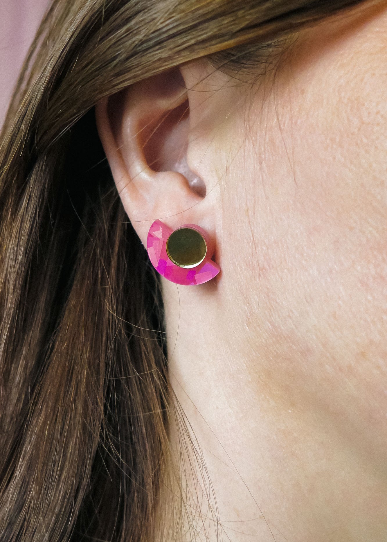 Close-up of a persons ear wearing lightweight Minimalist Geometric Stud Earrings, featuring a round gold stud and a pink, crescent-shaped accent. The person has straight brown hair tucked behind the ear.
