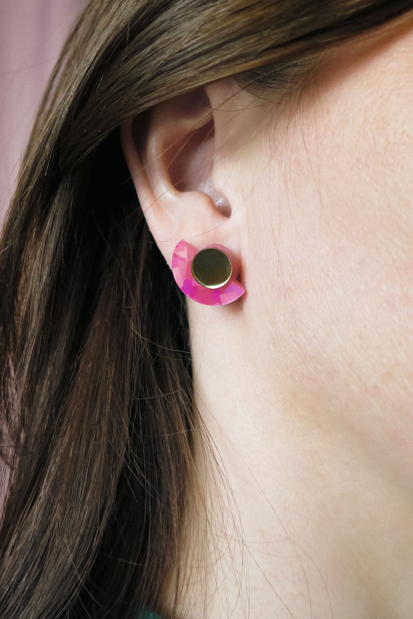 Close-up of a persons ear wearing lightweight Minimalist Geometric Stud Earrings, featuring a round gold stud and a pink, crescent-shaped accent. The person has straight brown hair tucked behind the ear.