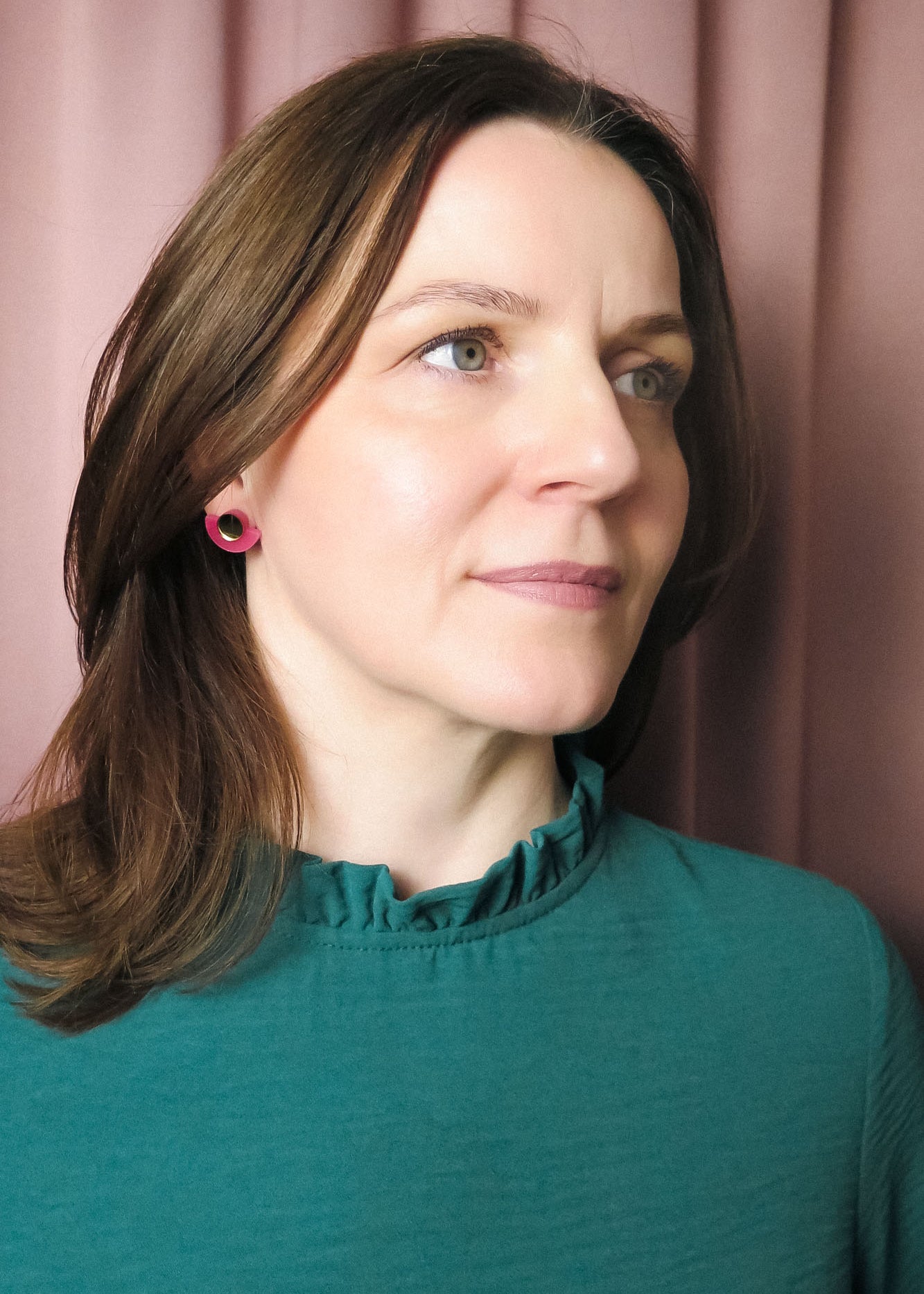 A woman with shoulder-length brown hair wearing a teal top with a ruffled collar and Minimalist Geometric Stud Earrings stands in front of pink curtains, looking slightly to the right with a neutral expression.