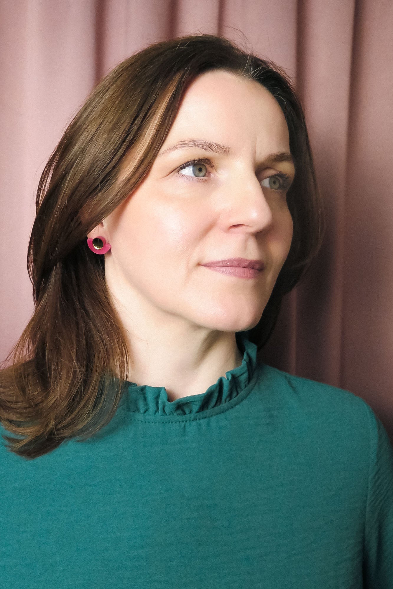 A woman with shoulder-length brown hair wearing a teal top with a ruffled collar and Minimalist Geometric Stud Earrings stands in front of pink curtains, looking slightly to the right with a neutral expression.