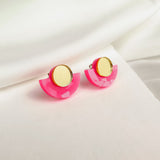Minimalist Geometric Stud Earrings featuring gold circular tops and bright pink, semi-circle bases are displayed on soft white fabric. These lightweight, hypoallergenic studs add a playful touch to any look.