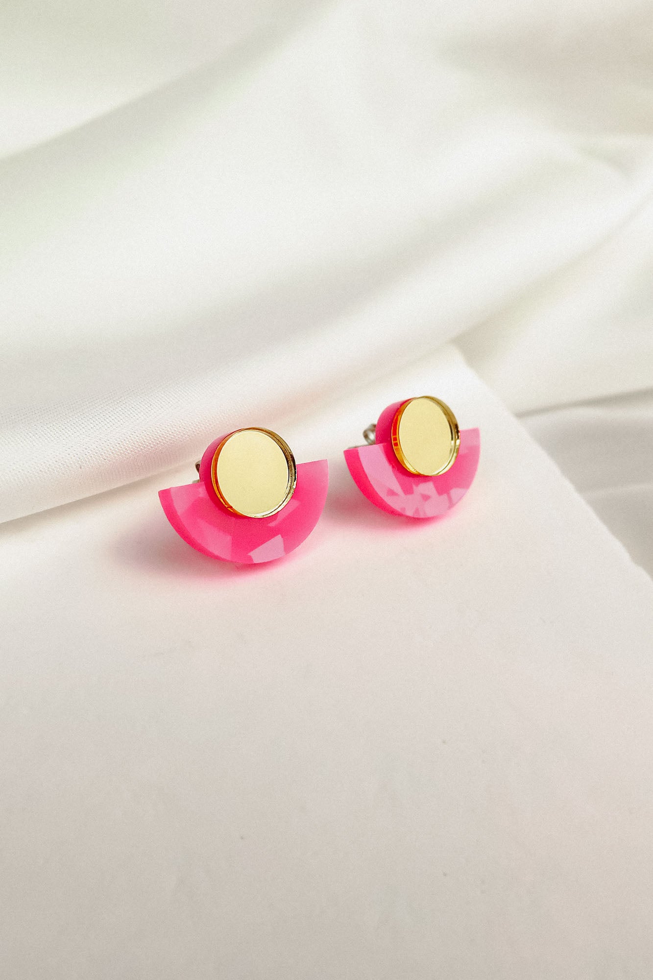 Minimalist Geometric Stud Earrings featuring gold circular tops and bright pink, semi-circle bases are displayed on soft white fabric. These lightweight, hypoallergenic studs add a playful touch to any look.