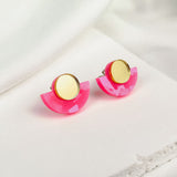 Minimalist Geometric Stud Earrings featuring gold circular tops and pink acrylic semicircles. These lightweight, hypoallergenic studs are displayed on a white fabric surface.