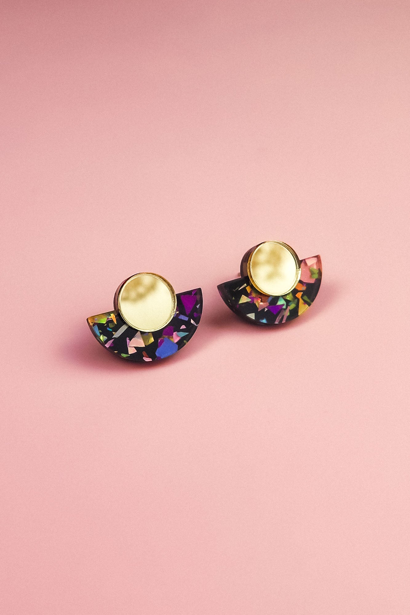 A pair of minimalist geometric stud earrings with gold circular tops and black semicircular bases featuring vibrant, multicolored confetti-like patterns, these hypoallergenic, lightweight pieces are displayed on a pink background.