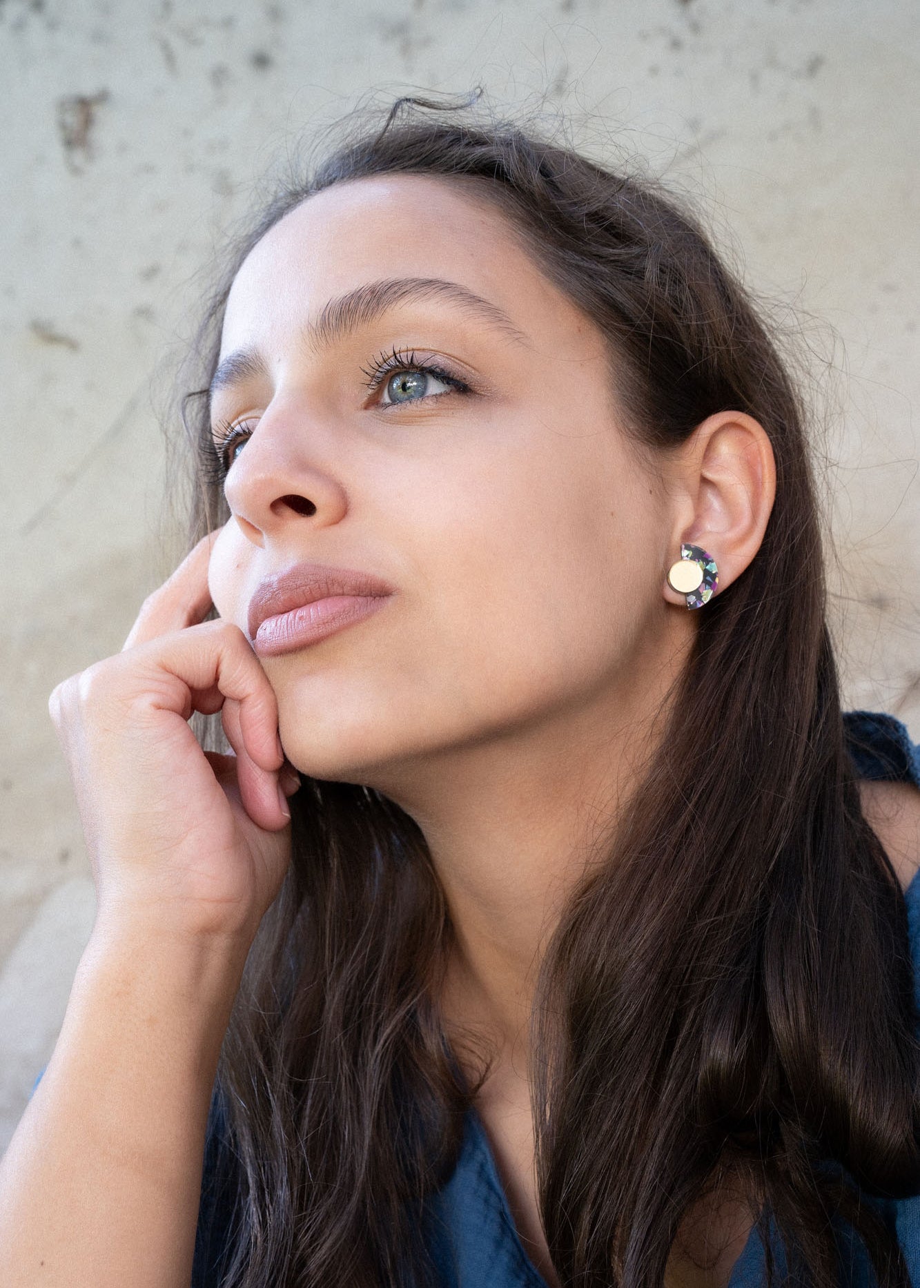 A woman with long brown hair and light eyes looks thoughtfully into the distance, resting her chin on her hand. She is wearing a blue top and minimalist geometric stud earrings, with a neutral, textured background behind her.