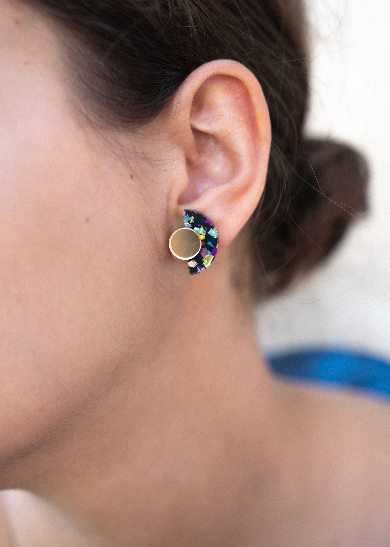 Close-up of a woman’s ear wearing colorful, crescent-shaped Minimalist Geometric Stud Earrings with a round gold stud. The womans dark hair is tied back in a bun. These hypoallergenic earrings are lightweight and set against a softly blurred background.
