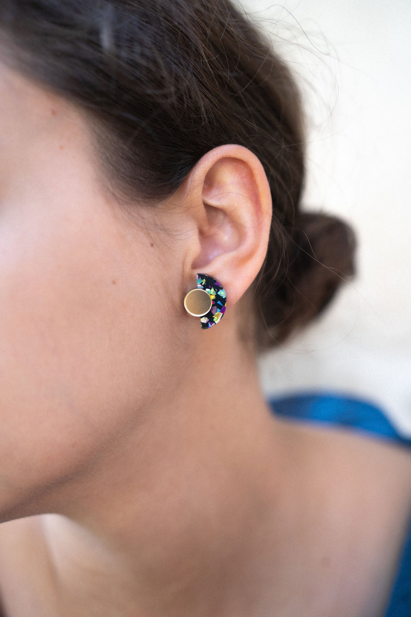 Close-up of a woman’s ear wearing colorful, crescent-shaped Minimalist Geometric Stud Earrings with a round gold stud. The womans dark hair is tied back in a bun. These hypoallergenic earrings are lightweight and set against a softly blurred background.