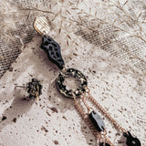 A modern, artistic mismatched extravagant earring in gold, black, and tortoiseshell rests on a textured stone surface, surrounded by dried foliage and beige woven fabric in soft natural light.