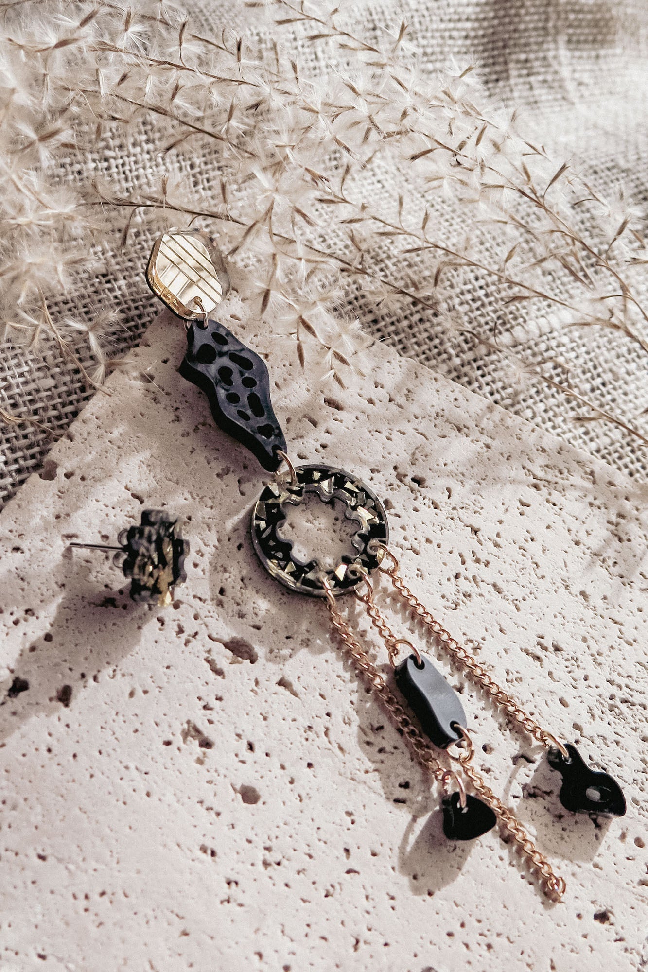 A modern, artistic mismatched extravagant earring in gold, black, and tortoiseshell rests on a textured stone surface, surrounded by dried foliage and beige woven fabric in soft natural light.