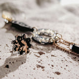 Close-up of a stylish gold bracelet and a matching floral-shaped ring with black and gold details, paired with lightweight handcrafted earrings, all arranged on a textured, light-colored stone surface in soft natural light.