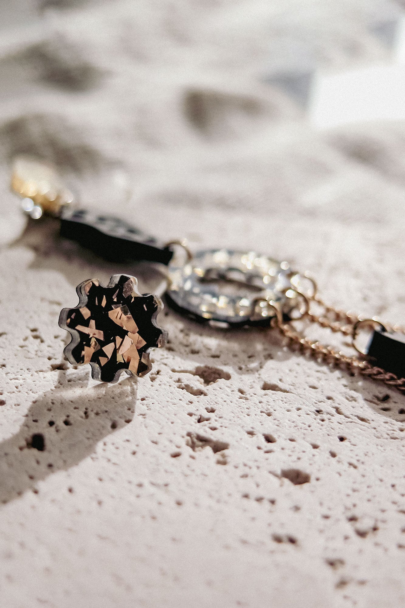 Close-up of a stylish gold bracelet and a matching floral-shaped ring with black and gold details, paired with lightweight handcrafted earrings, all arranged on a textured, light-colored stone surface in soft natural light.