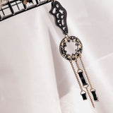 A single handcrafted earring with geometric shapes in gold and black, hanging from a metal rack. This lightweight earring features abstract and circular elements with dangling chains and small black accents.