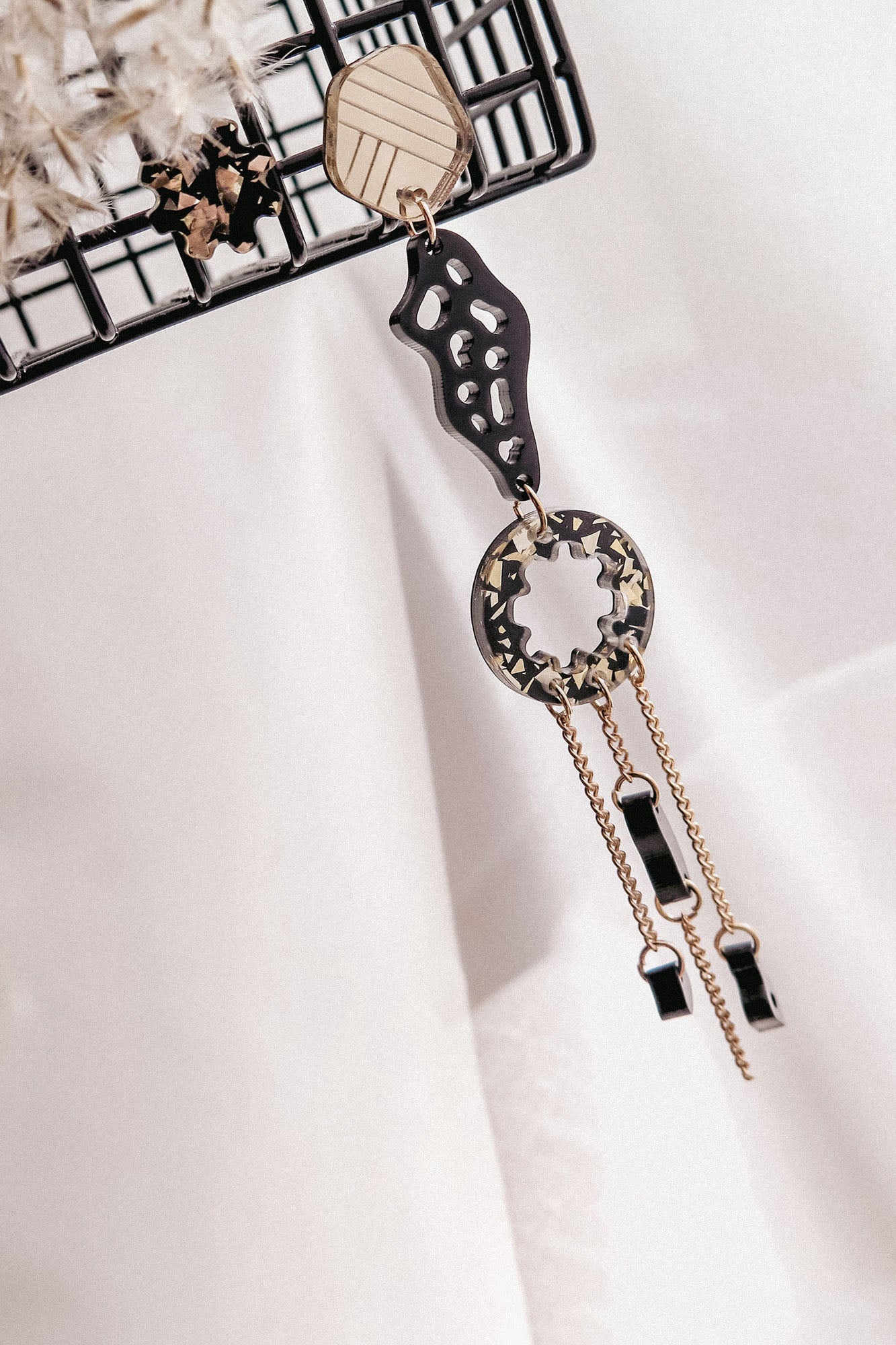 A single handcrafted earring with geometric shapes in gold and black, hanging from a metal rack. This lightweight earring features abstract and circular elements with dangling chains and small black accents.