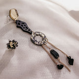 A single handcrafted statement earring with geometric shapes, black and gold accents, and dangling chains lies on textured white fabric. Another small, round black and gold earring is placed nearby for a mismatched extravagant look.