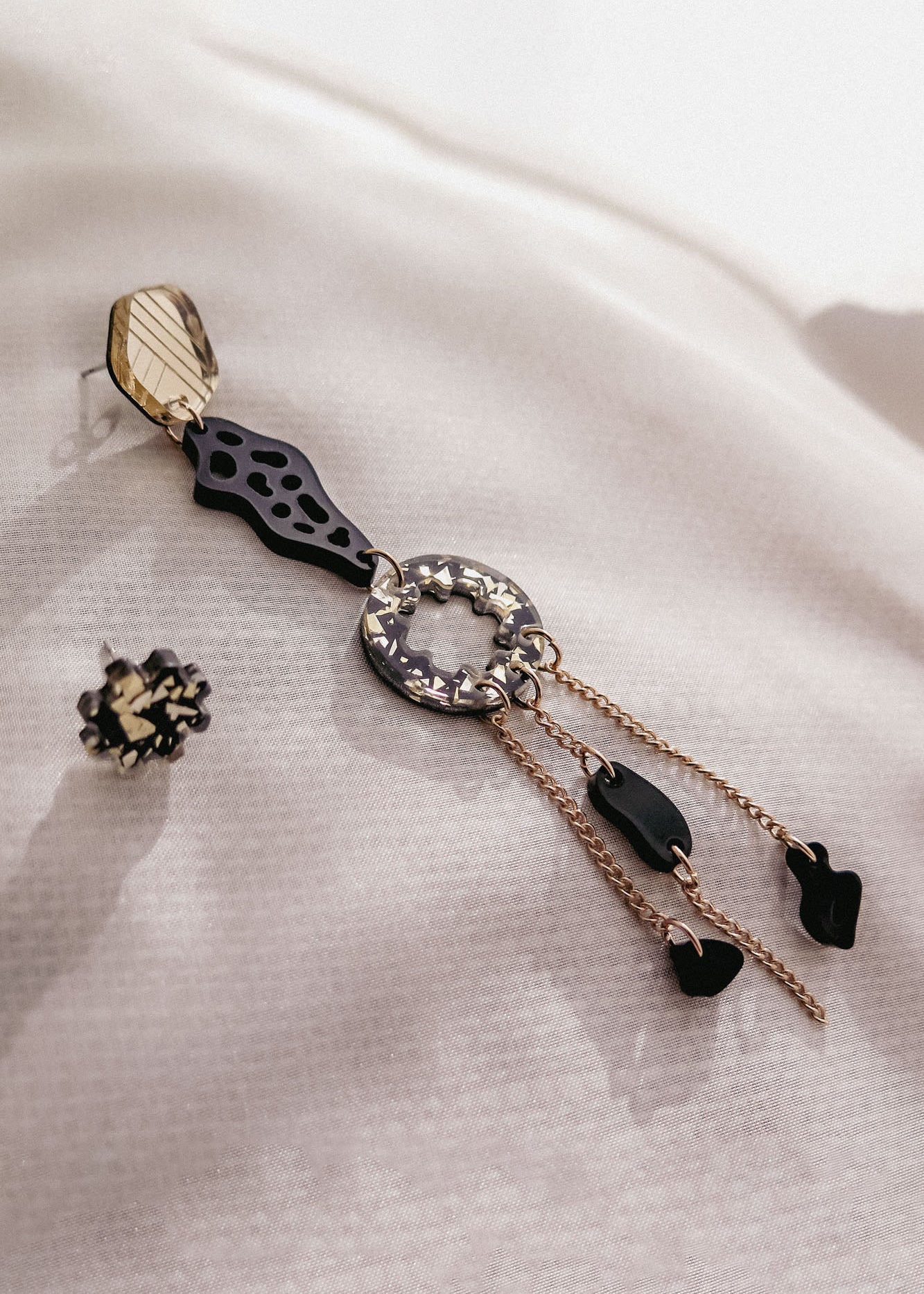 A single handcrafted statement earring with geometric shapes, black and gold accents, and dangling chains lies on textured white fabric. Another small, round black and gold earring is placed nearby for a mismatched extravagant look.