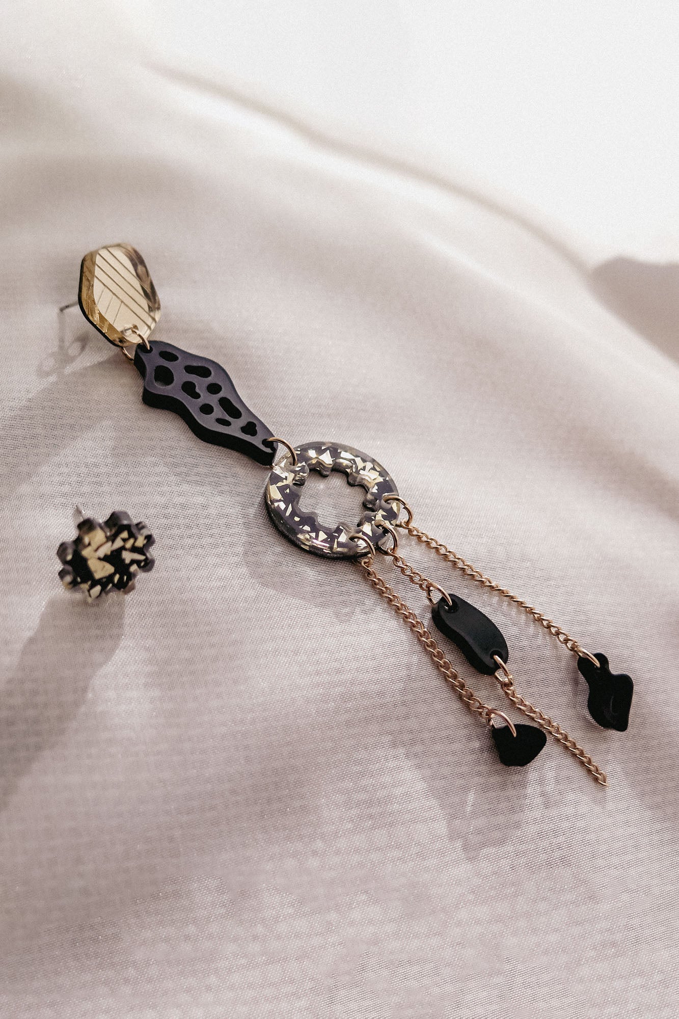 A single handcrafted statement earring with geometric shapes, black and gold accents, and dangling chains lies on textured white fabric. Another small, round black and gold earring is placed nearby for a mismatched extravagant look.