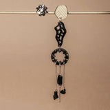 A single handcrafted earring featuring black and silver geometric shapes and dangling chains hangs from a horizontal metallic rod against a beige background—perfect for those who love lightweight earrings with an abstract flair.