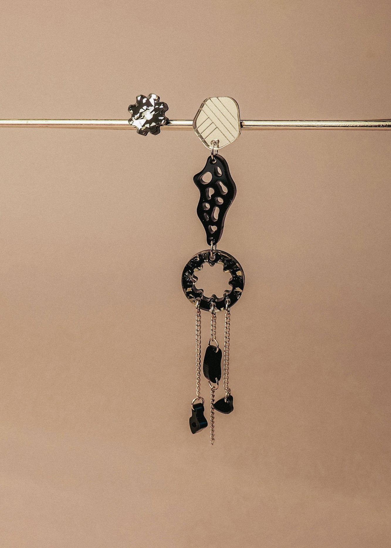 A single handcrafted earring featuring black and silver geometric shapes and dangling chains hangs from a horizontal metallic rod against a beige background—perfect for those who love lightweight earrings with an abstract flair.