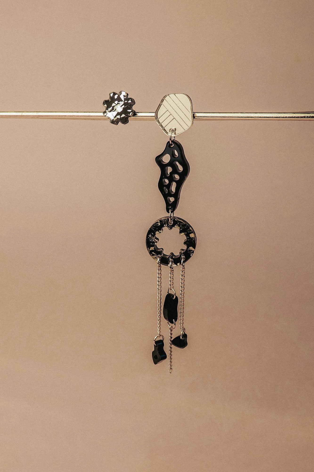 A single handcrafted earring featuring black and silver geometric shapes and dangling chains hangs from a horizontal metallic rod against a beige background—perfect for those who love lightweight earrings with an abstract flair.