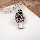 A handcrafted Morel Mushroom Pin, made from eco-friendly materials, features a textured brown cap and white stem. This decorative enamel pin is displayed on a white surface with dried grass in the blurred background.