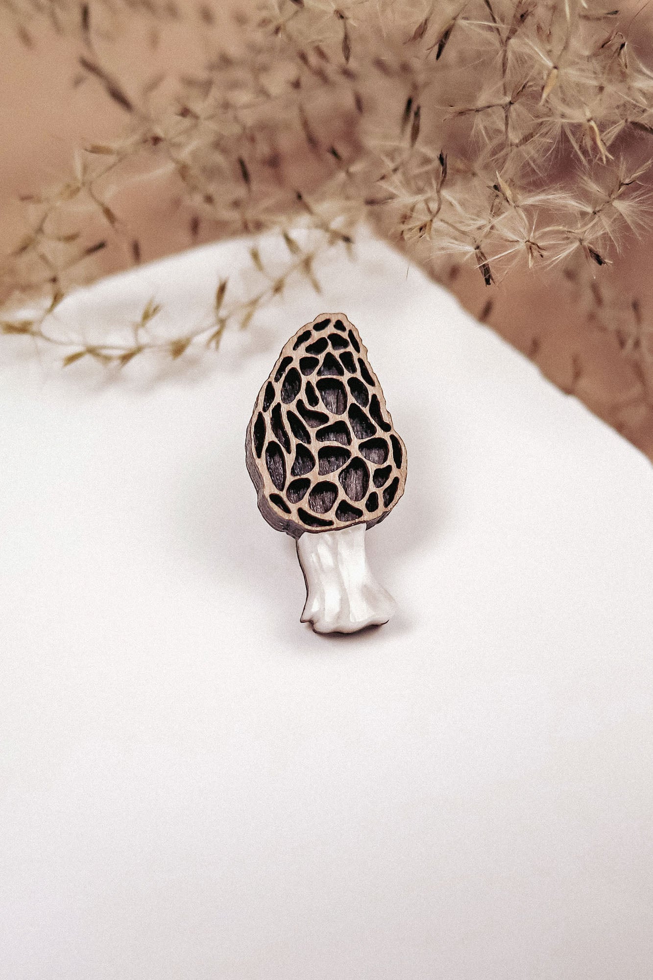 A handcrafted Morel Mushroom Pin, made from eco-friendly materials, features a textured brown cap and white stem. This decorative enamel pin is displayed on a white surface with dried grass in the blurred background.