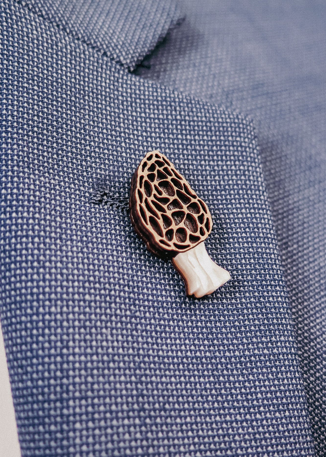 A close-up of a blue textured blazer with a handcrafted Morel Mushroom Pin, made from eco-friendly materials, attached to its lapel.