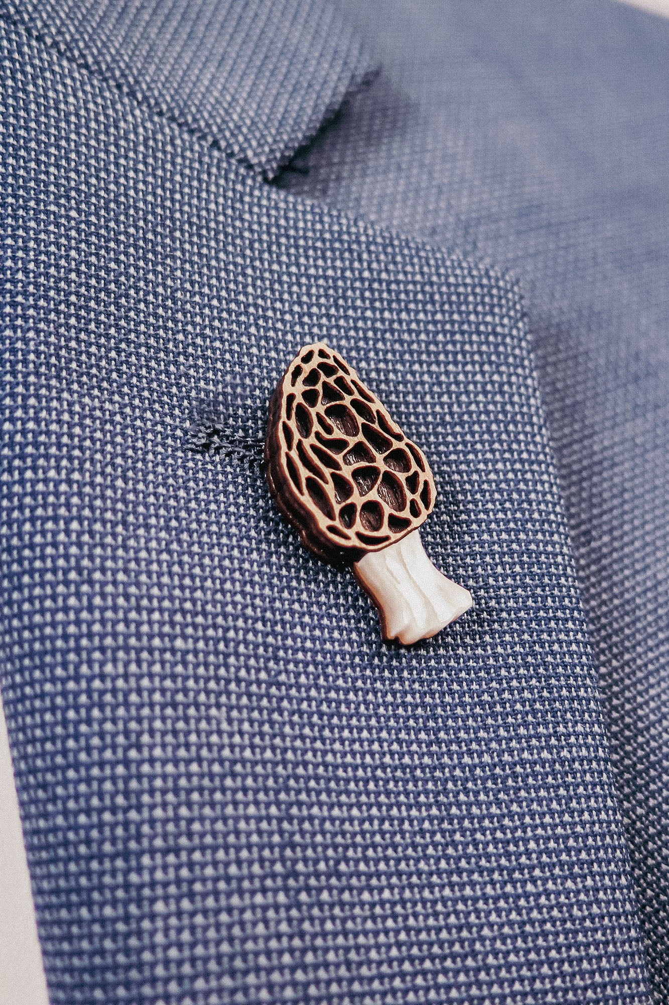 A close-up of a blue textured blazer with a handcrafted Morel Mushroom Pin, made from eco-friendly materials, attached to its lapel.