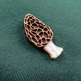 A small, detailed Morel Mushroom Pin, handcrafted from eco-friendly materials, rests on a green textured fabric background.
