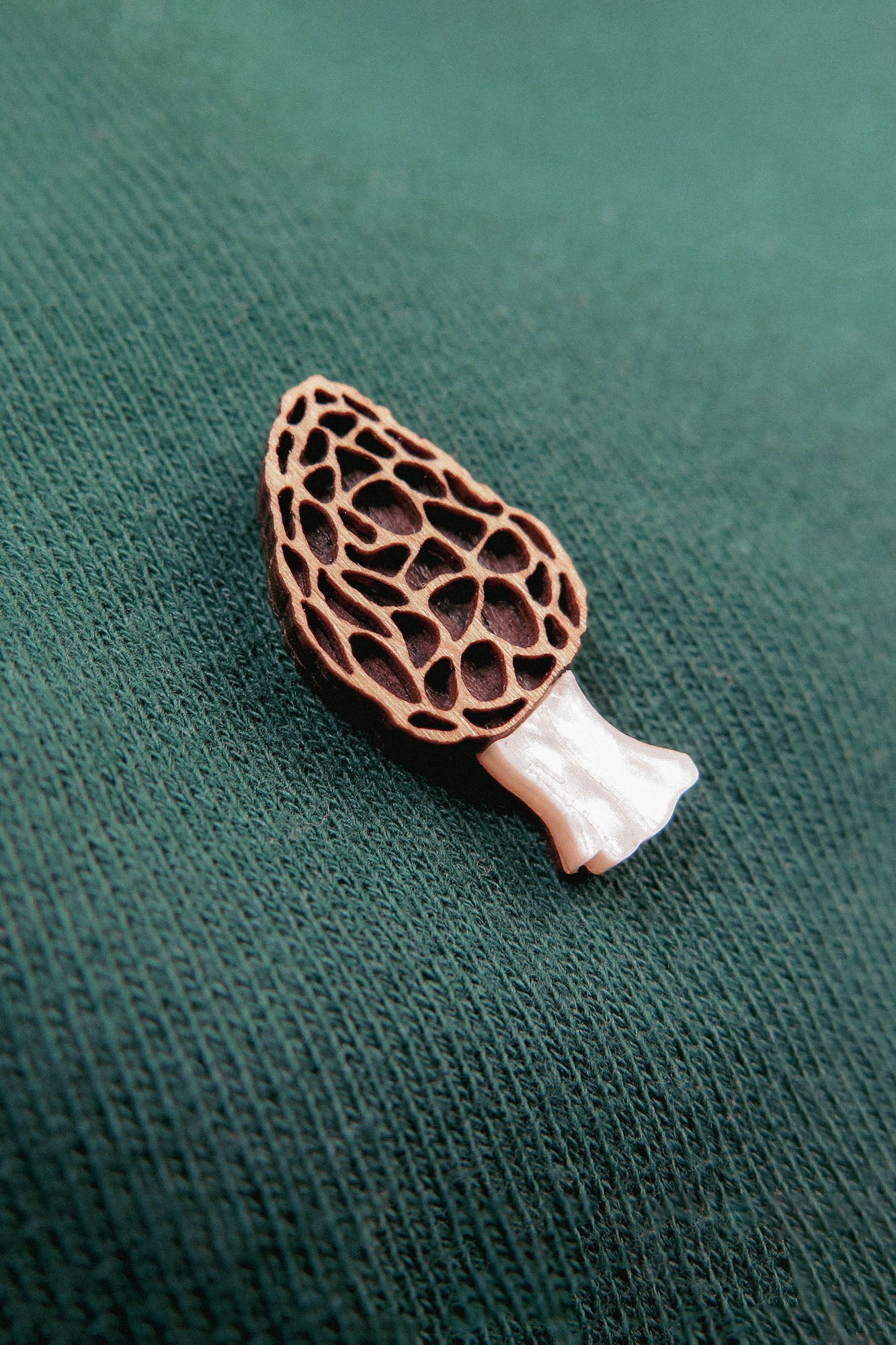 A small, detailed Morel Mushroom Pin, handcrafted from eco-friendly materials, rests on a green textured fabric background.