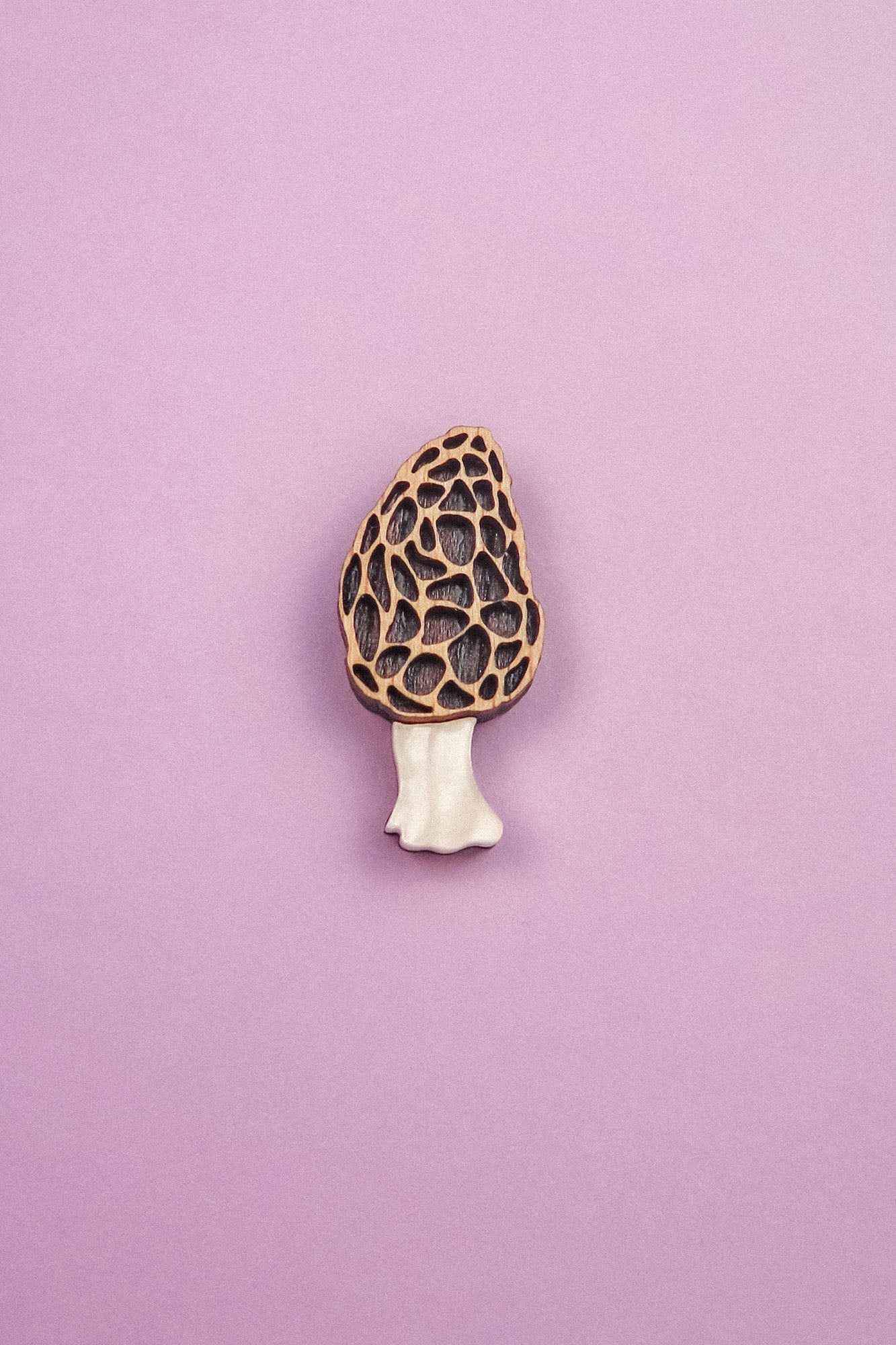 A handcrafted Morel Mushroom Pin made from eco-friendly materials features a textured, honeycomb-patterned cap and pale stem, displayed on a light purple background.