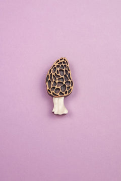 A handcrafted Morel Mushroom Pin made from eco-friendly materials features a textured, honeycomb-patterned cap and pale stem, displayed on a light purple background.