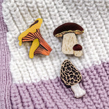 Three decorative mushroom pins, handcrafted from eco-friendly materials—including a Morel Mushroom Pin with a honeycomb pattern—are displayed on a knitted blanket with white and lavender stripes.