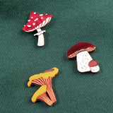 Three colorful, handcrafted enamel pins shaped like different mushrooms—including a Morel Mushroom Pin—are attached to a green fabric surface. These vibrant pins are made using eco-friendly materials for a sustainable touch.