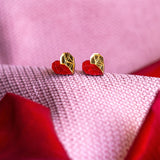 Gold and red geometric heart studs rest on pink and red textured fabric, with the colors and patterns creating a soft, romantic background—a perfect touch of romantic jewelry.