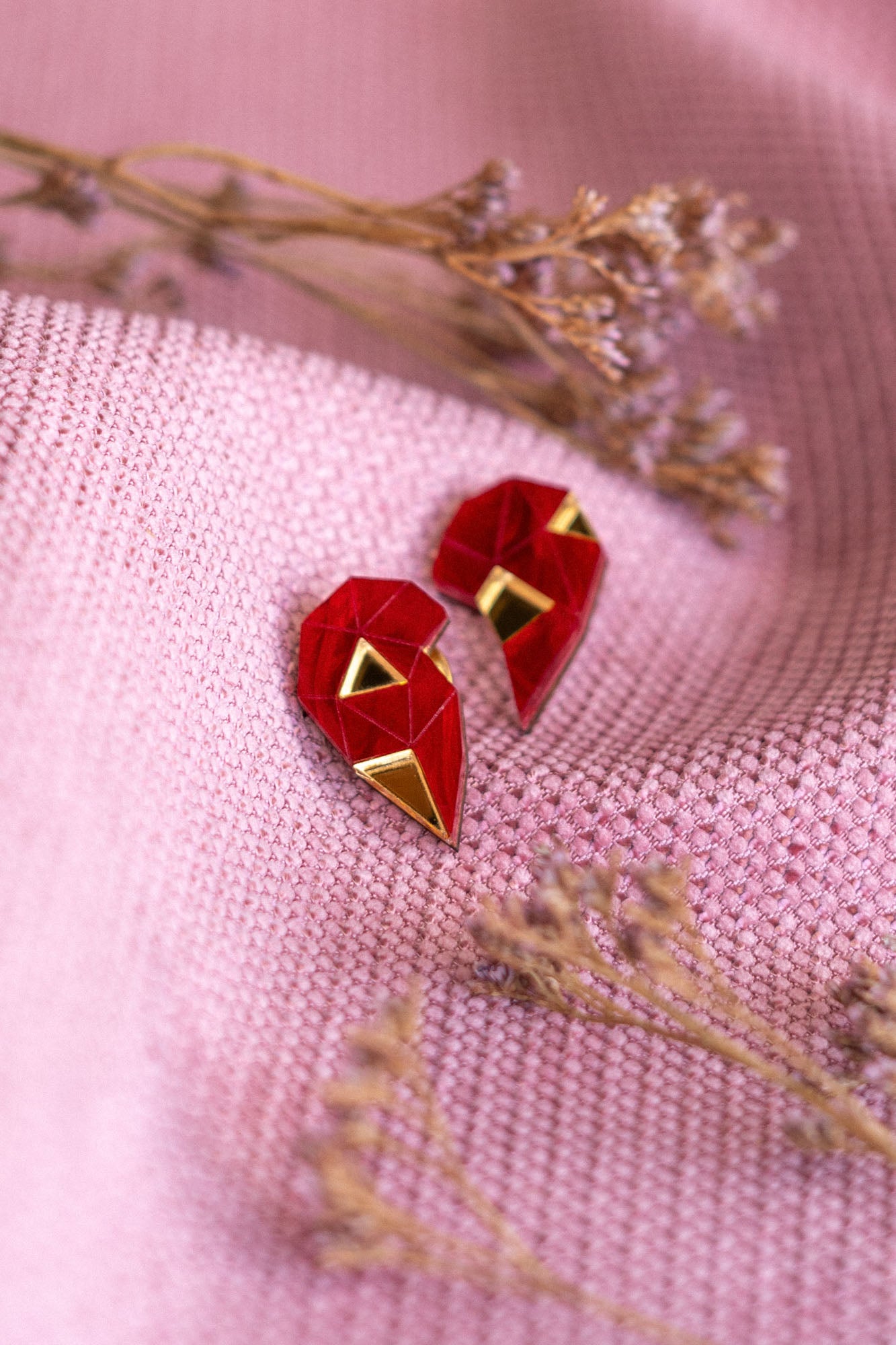 A pair of handcrafted geometric red and gold earrings shaped like hearts rest on textured pink fabric, with small dried flowers placed nearby—a perfect romantic gift.