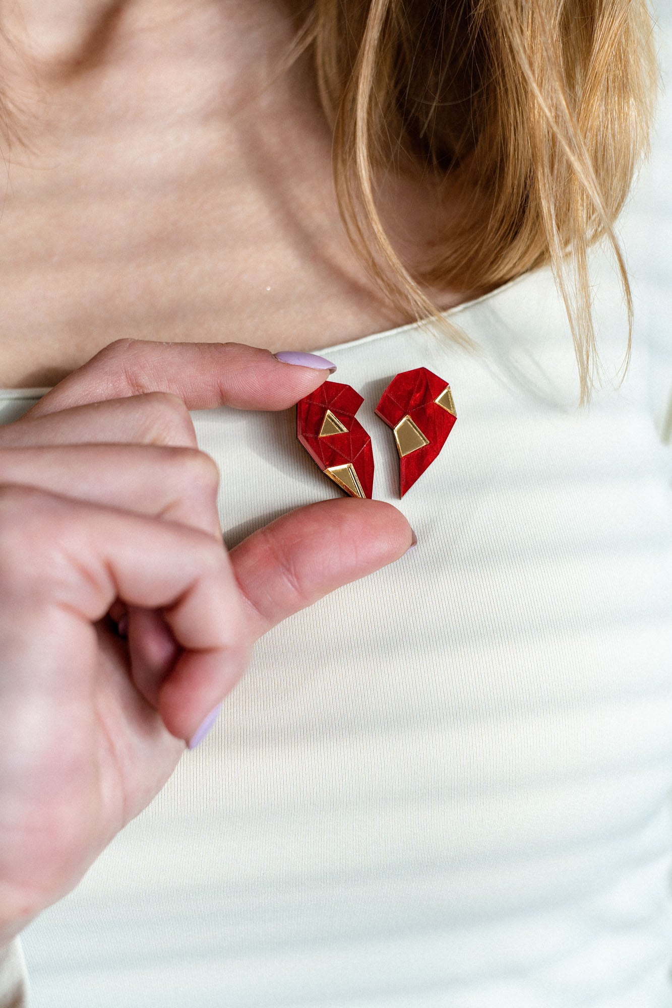 A person wearing a white top holds a red, geometric, handcrafted One Love Halves Pin against their chest. The heart-shaped pin is split into two pieces with gold accents, making it a meaningful romantic gift.