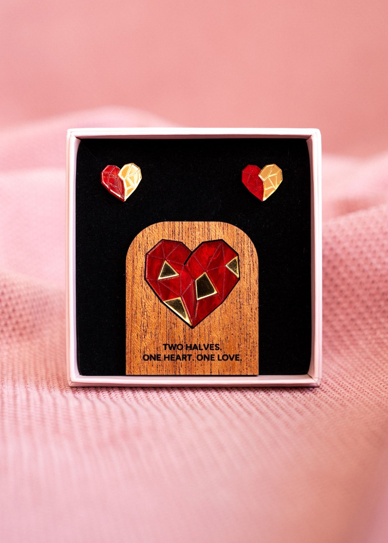 A jewelry set with geometric polygon hearts earrings and a matching pin displayed in a box. The pin features a red and gold heart on wood with the text: TWO HALVES. ONE HEART. ONE LOVE.—the perfect Valentine’s statement. Soft pink fabric background.