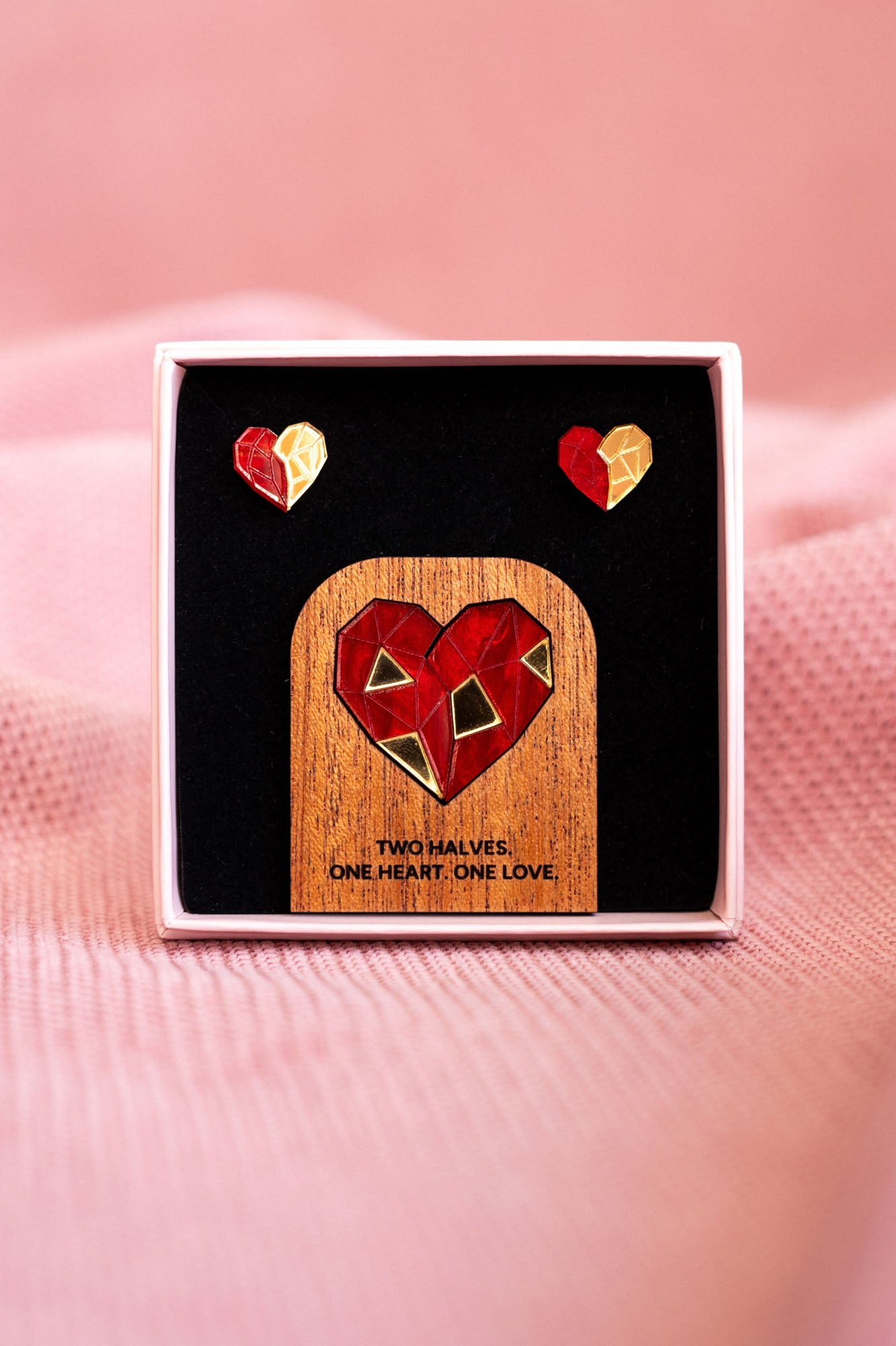 A jewelry set with geometric polygon hearts earrings and a matching pin displayed in a box. The pin features a red and gold heart on wood with the text: TWO HALVES. ONE HEART. ONE LOVE.—the perfect Valentine’s statement. Soft pink fabric background.