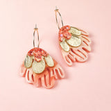 A pair of gold hoops with layered pink acrylic petals, gold-toned metal accents, and pink sequins evoke the charm of Organic Floral Hoops, beautifully arranged on a light pink background.