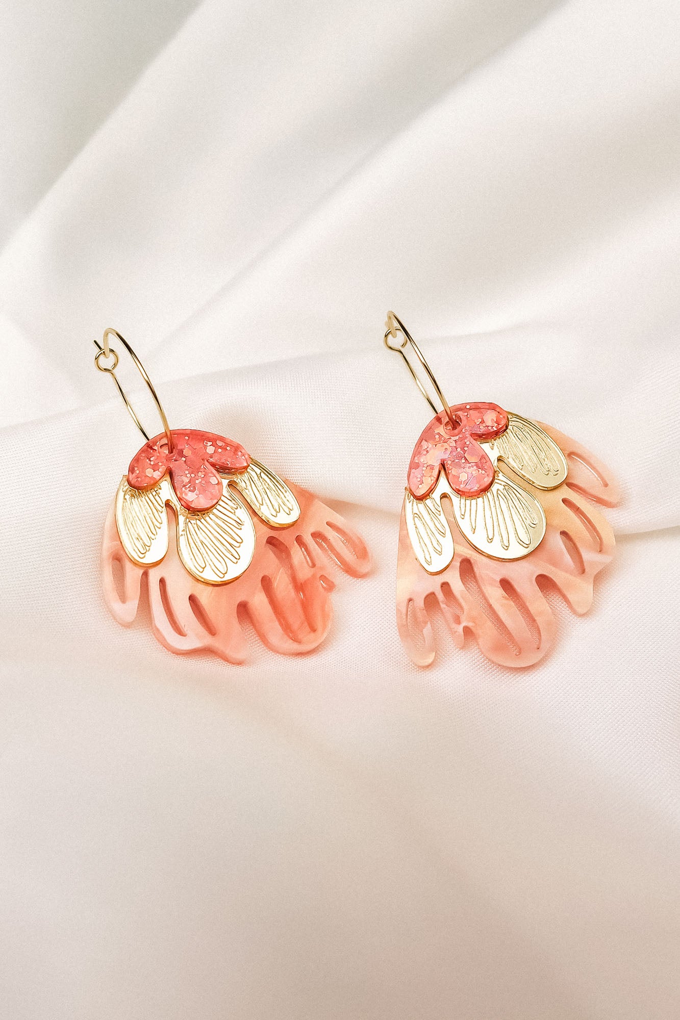 A pair of decorative Organic Floral Hoops featuring pink translucent petal-like acrylic flowers and gold accents, displayed on a soft white fabric background.
