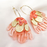 A pair of Organic Floral Hoops with pink, coral, and gold petal-like acrylic flowers rest on a soft white fabric background. These gold hoops feature intricate cut-out designs and shimmering accents for a delicate, nature-inspired look.
