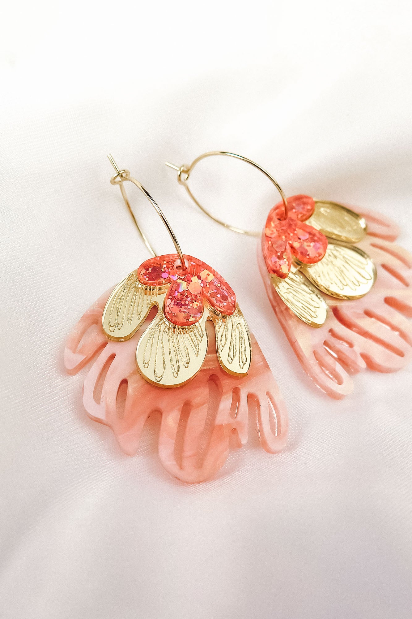 A pair of Organic Floral Hoops with pink, coral, and gold petal-like acrylic flowers rest on a soft white fabric background. These gold hoops feature intricate cut-out designs and shimmering accents for a delicate, nature-inspired look.