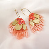 A pair of Organic Floral Hoops featuring gold, petal-shaped pieces and pink, coral-colored acrylic flowers, arranged in a floral design, displayed on a soft white fabric background.