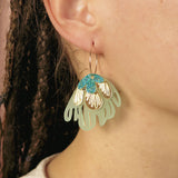 Close-up of a person’s ear adorned with handcrafted Organic Floral Hoops, large decorative earrings shaped like a flower in mint green, white, and blue. The person has braided hair and wears a light-colored top, radiating tropical elegance.