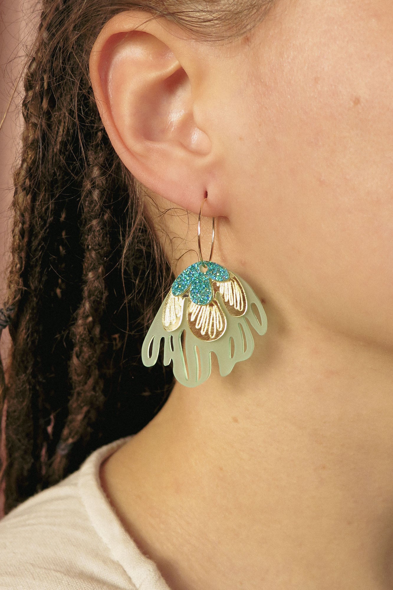 Close-up of a person’s ear adorned with handcrafted Organic Floral Hoops, large decorative earrings shaped like a flower in mint green, white, and blue. The person has braided hair and wears a light-colored top, radiating tropical elegance.