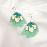 A pair of handcrafted dangling earrings with gold hooks, featuring layered designs in mint green, cream, and teal glitter, resembling abstract floral shapes for a touch of tropical elegance, displayed on a white fabric background.