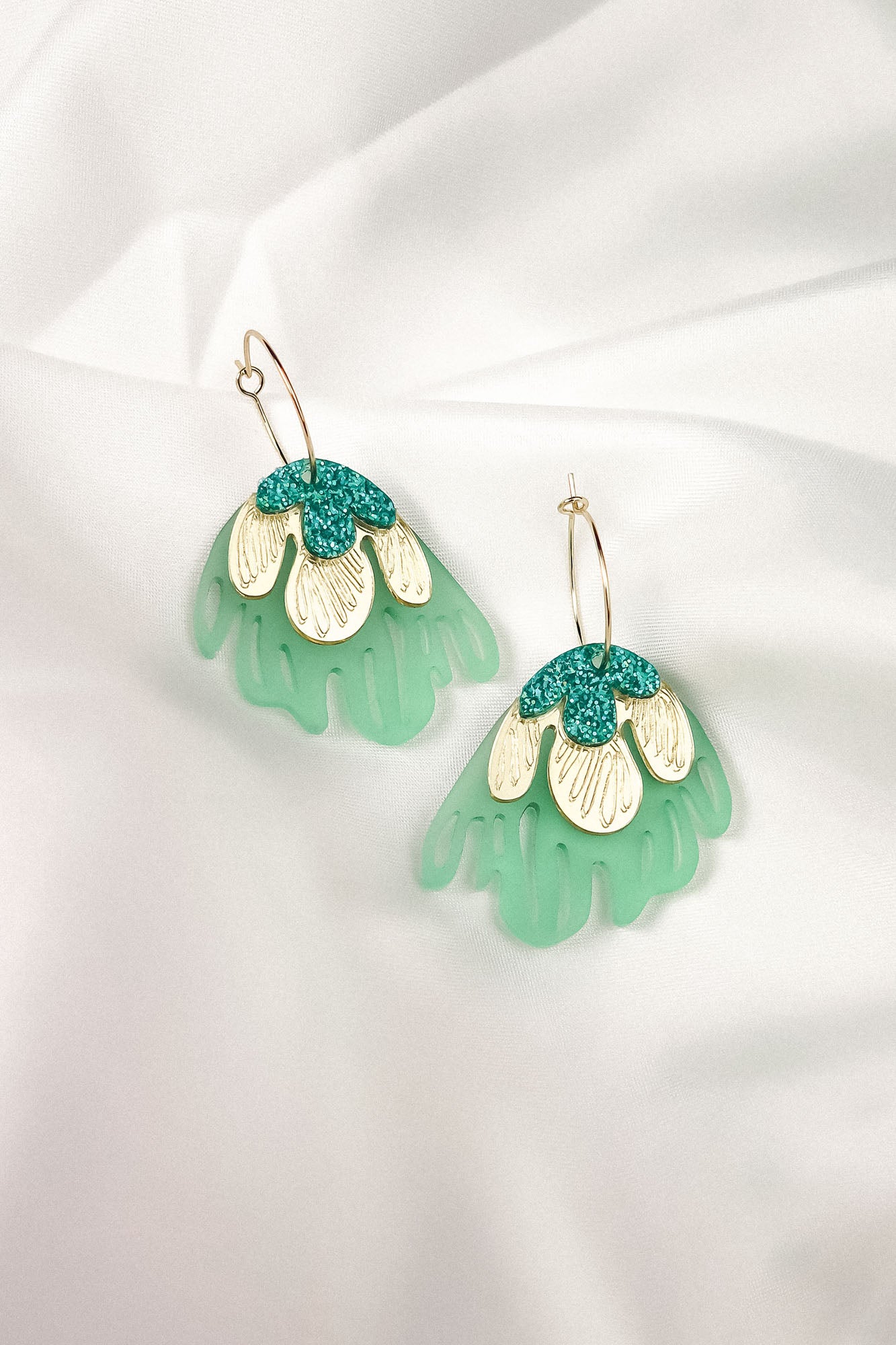 A pair of handcrafted dangling earrings with gold hooks, featuring layered designs in mint green, cream, and teal glitter, resembling abstract floral shapes for a touch of tropical elegance, displayed on a white fabric background.