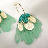 A close-up of a handcrafted gold hoop earring featuring green and gold petal-shaped charms with glittery accents, embodying tropical elegance, set on a soft white fabric background.