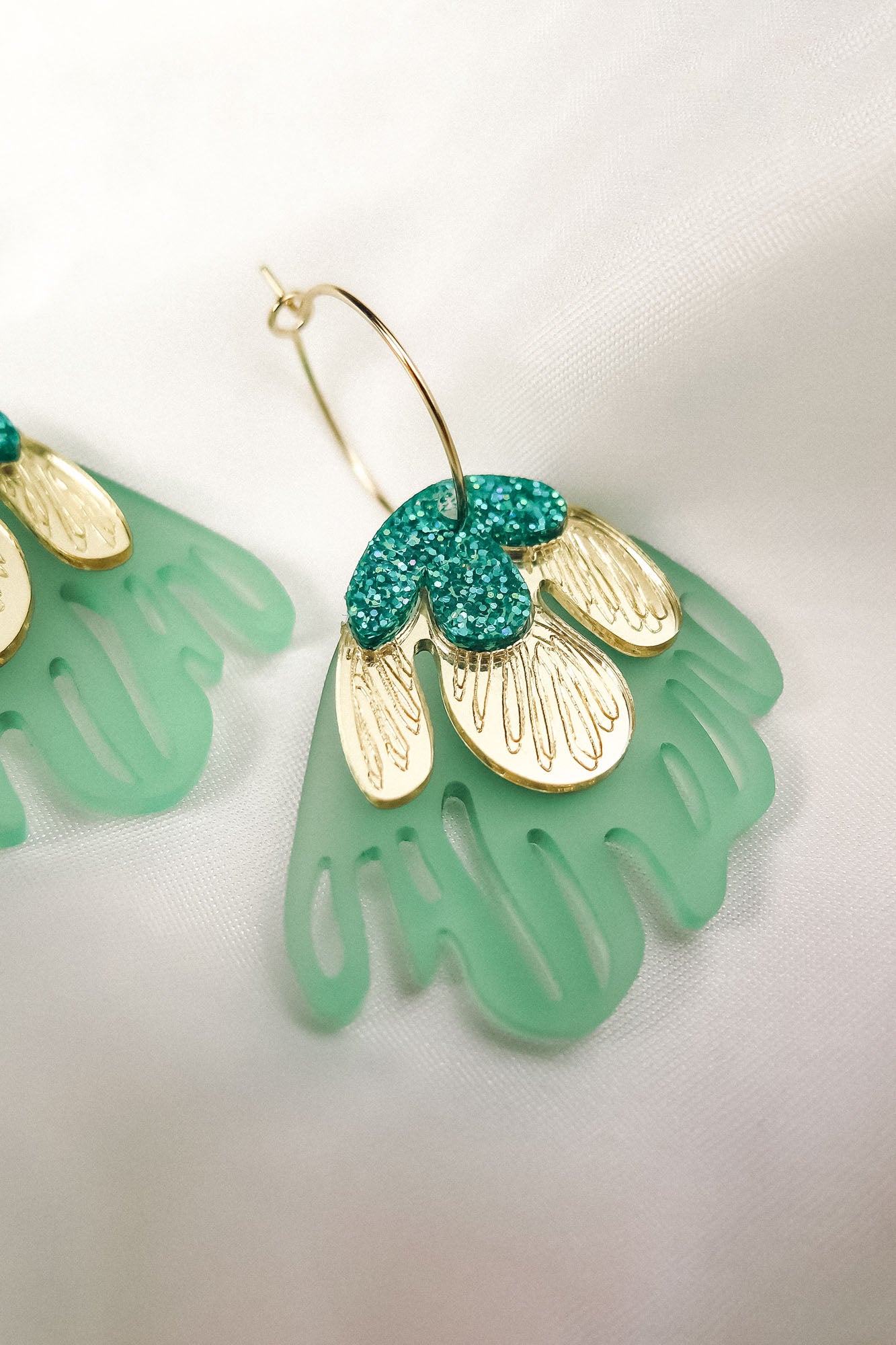A close-up of a handcrafted gold hoop earring featuring green and gold petal-shaped charms with glittery accents, embodying tropical elegance, set on a soft white fabric background.
