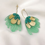 A pair of handcrafted earrings with gold metallic petals, glittery green accents, and translucent green acrylic shapes resembling leaves, displayed on white satin fabric—perfect for adding a touch of tropical elegance.